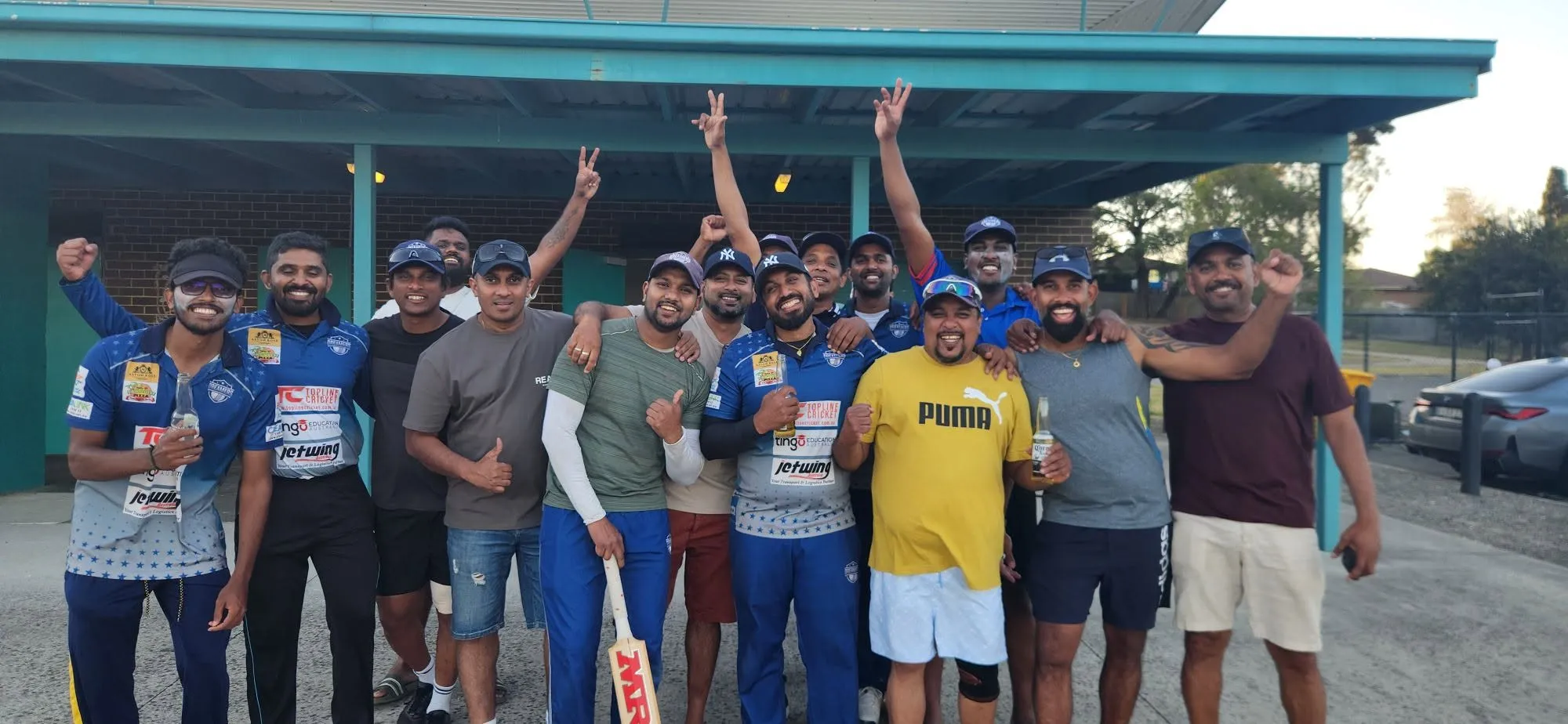The Dreamers squad with arms raised and bats held up, celebrating a win in front of the ground shelter.
