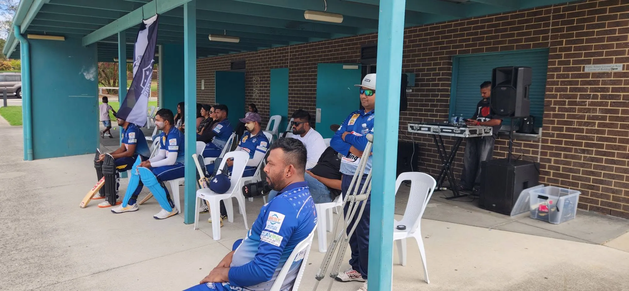 Players and supporters seated under the ground shelter on a match day, club flag flying beside them.