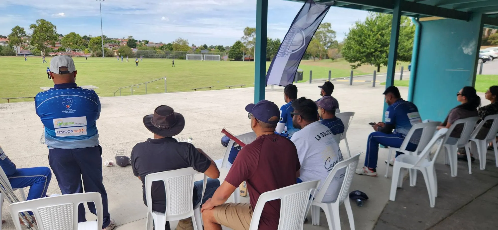 Players and supporters watching a match from the shelter, with the ground stretching out behind them.